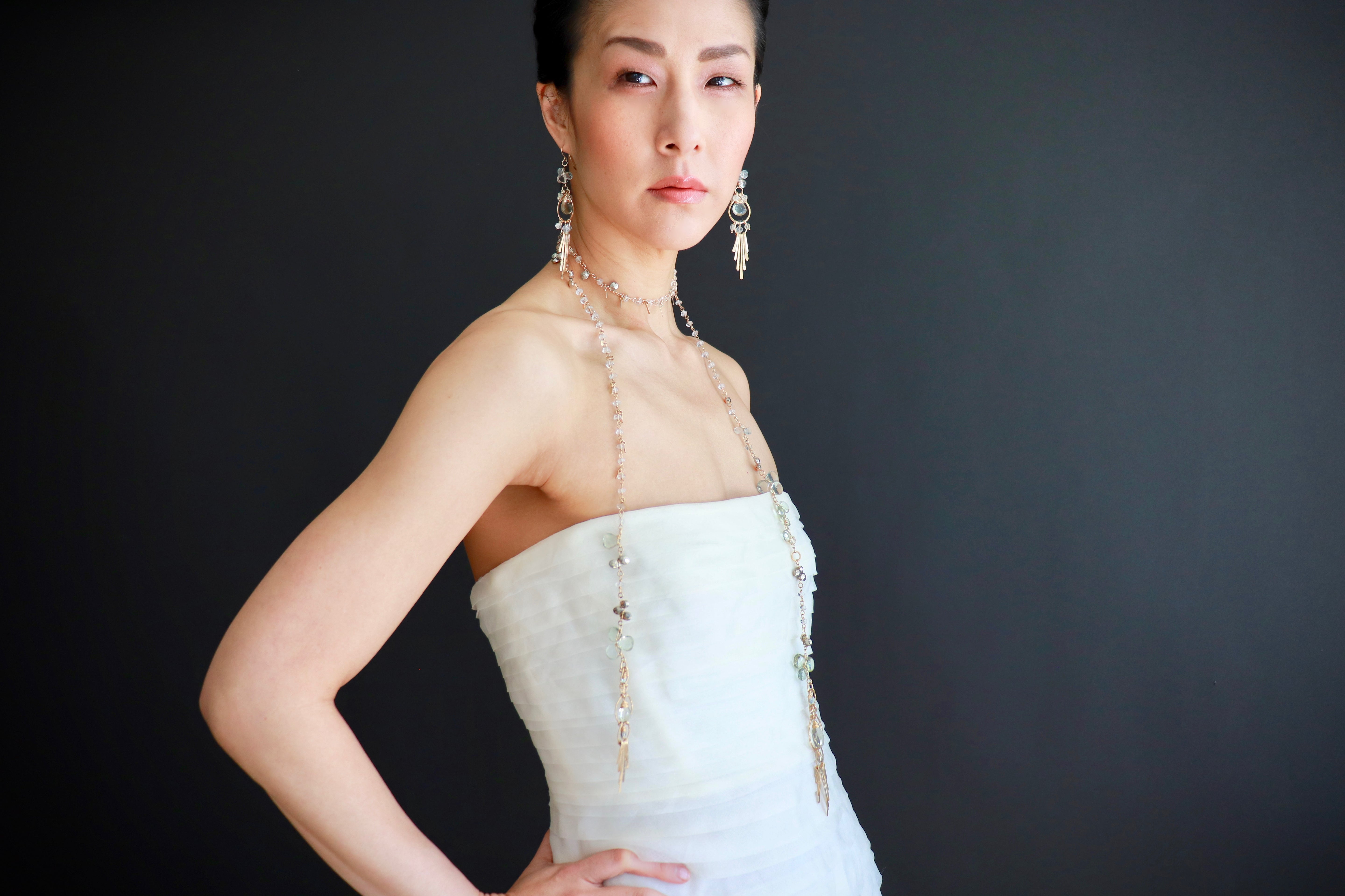 A woman wearing a strapless white dress and large earrings poses confidently against a dark background.