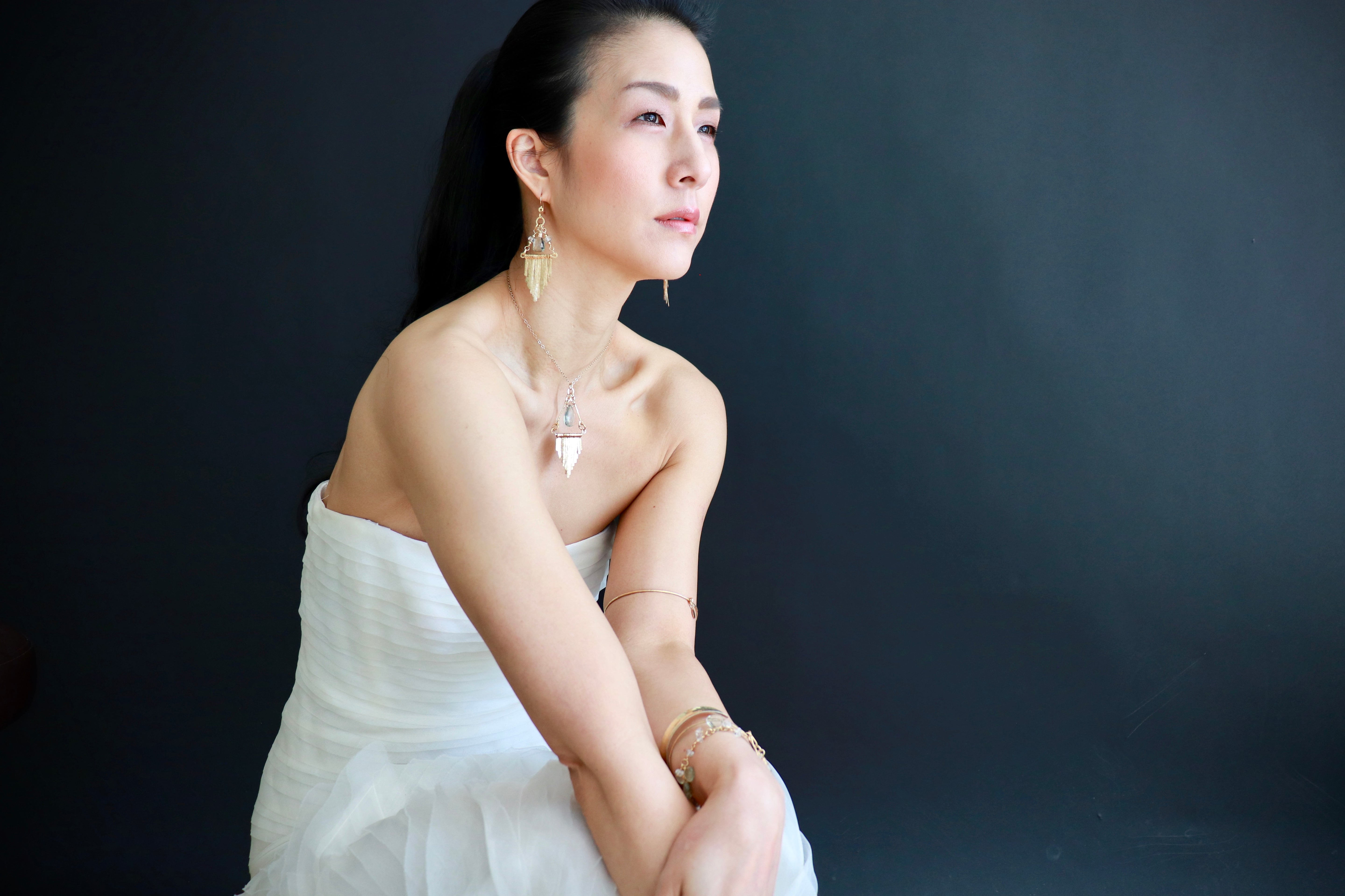 A woman in a white dress poses against a dark background, her gaze directed off to the side.