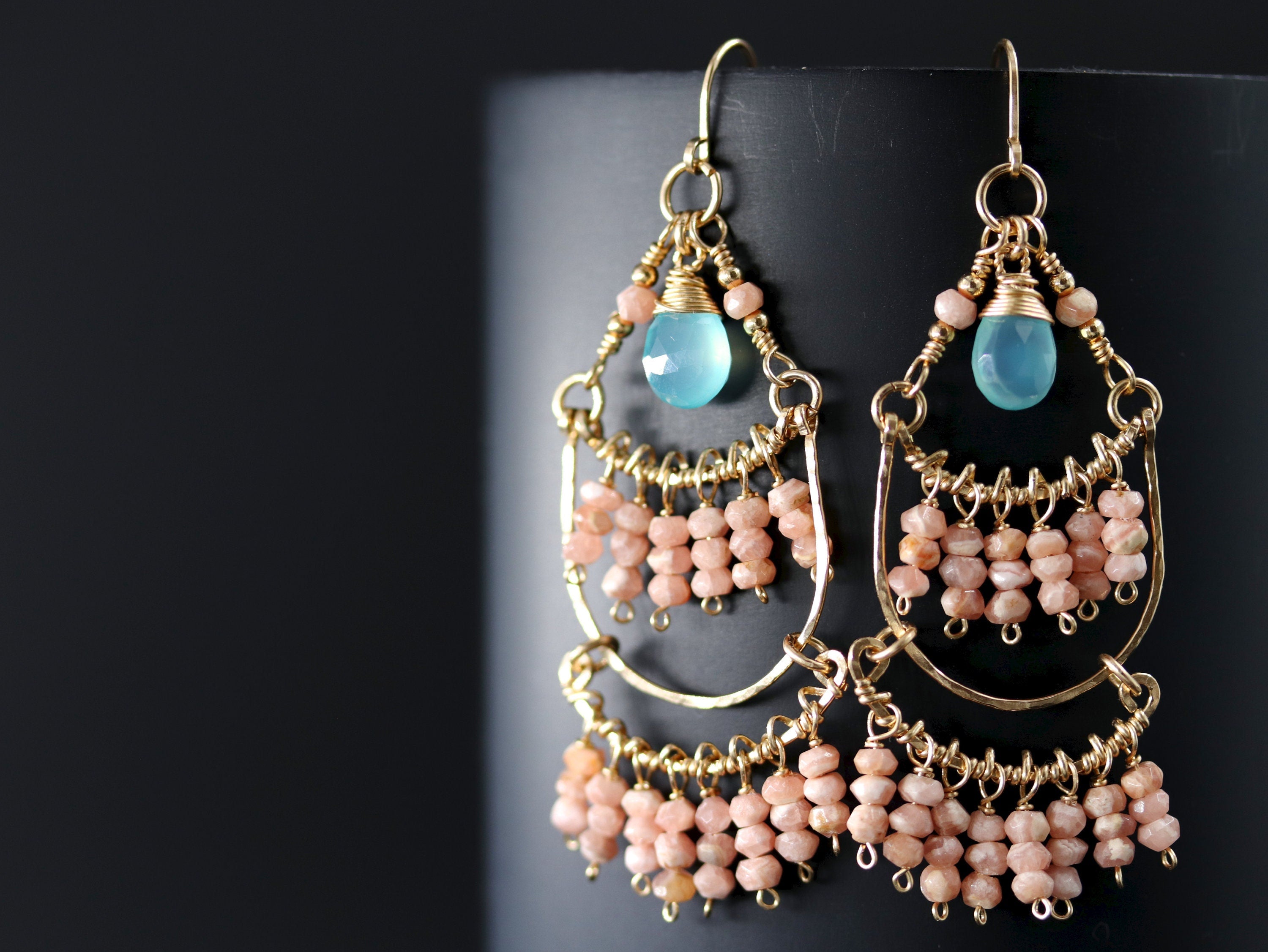 May include: Gold dangle earrings with light blue and peach-colored stones. The earrings have a layered design with multiple tiers of beads.