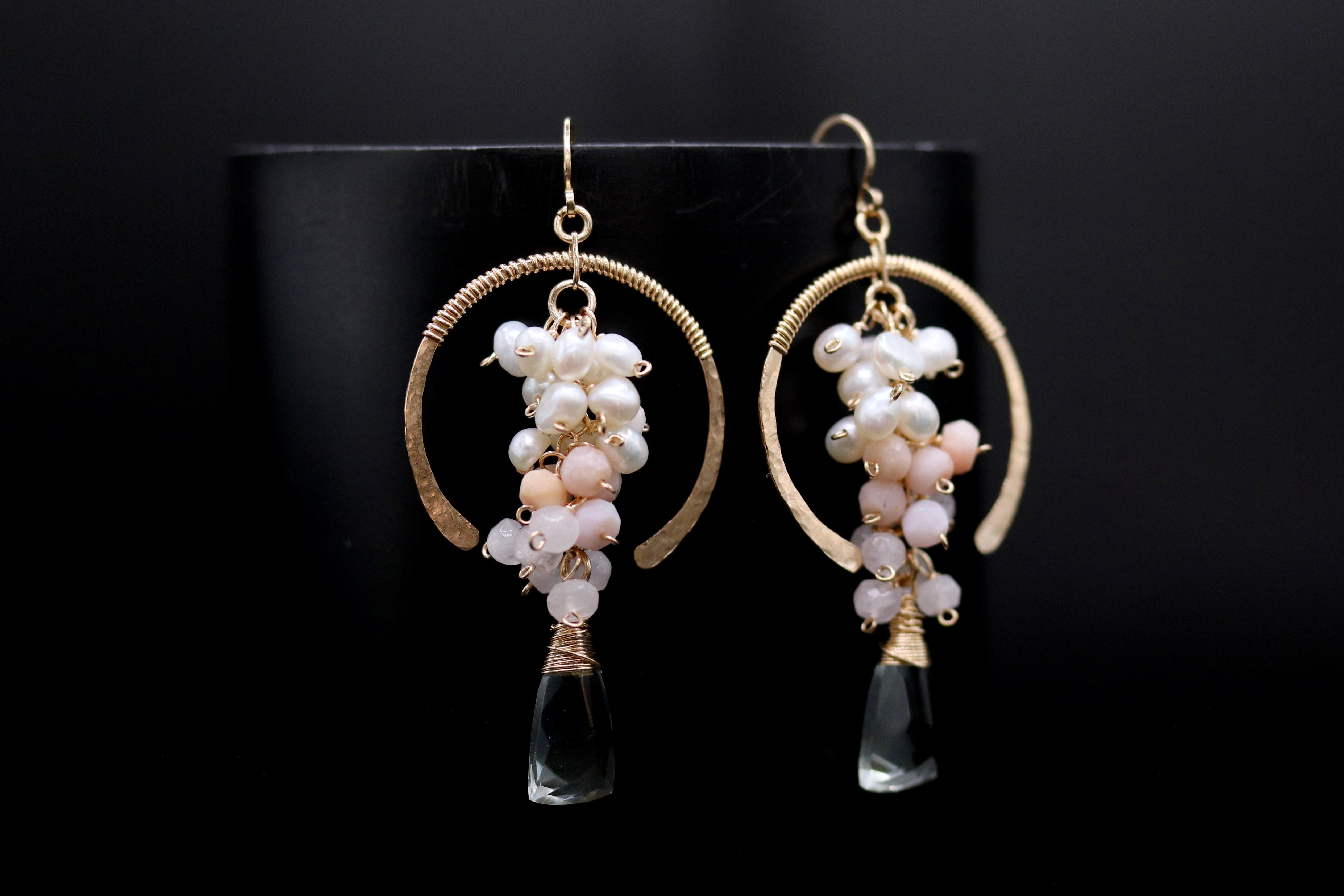 A pair of elegant earrings with a black background. The earrings have a gold hoop design and are adorned with small white and pink pearls, as well as clear gemstone accents.