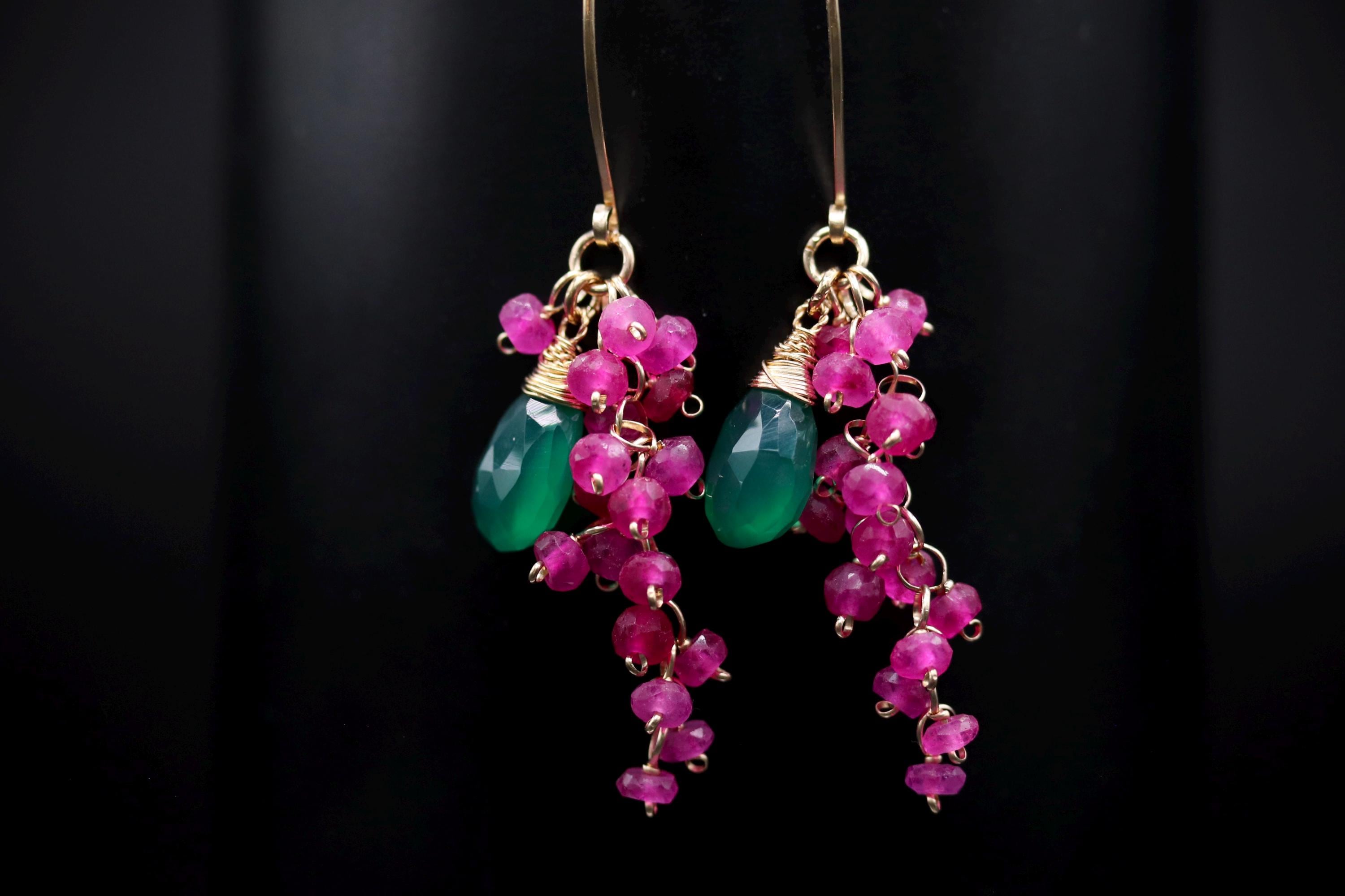 A pair of earrings with green and pink gemstones and dangling charms, set against a dark background.