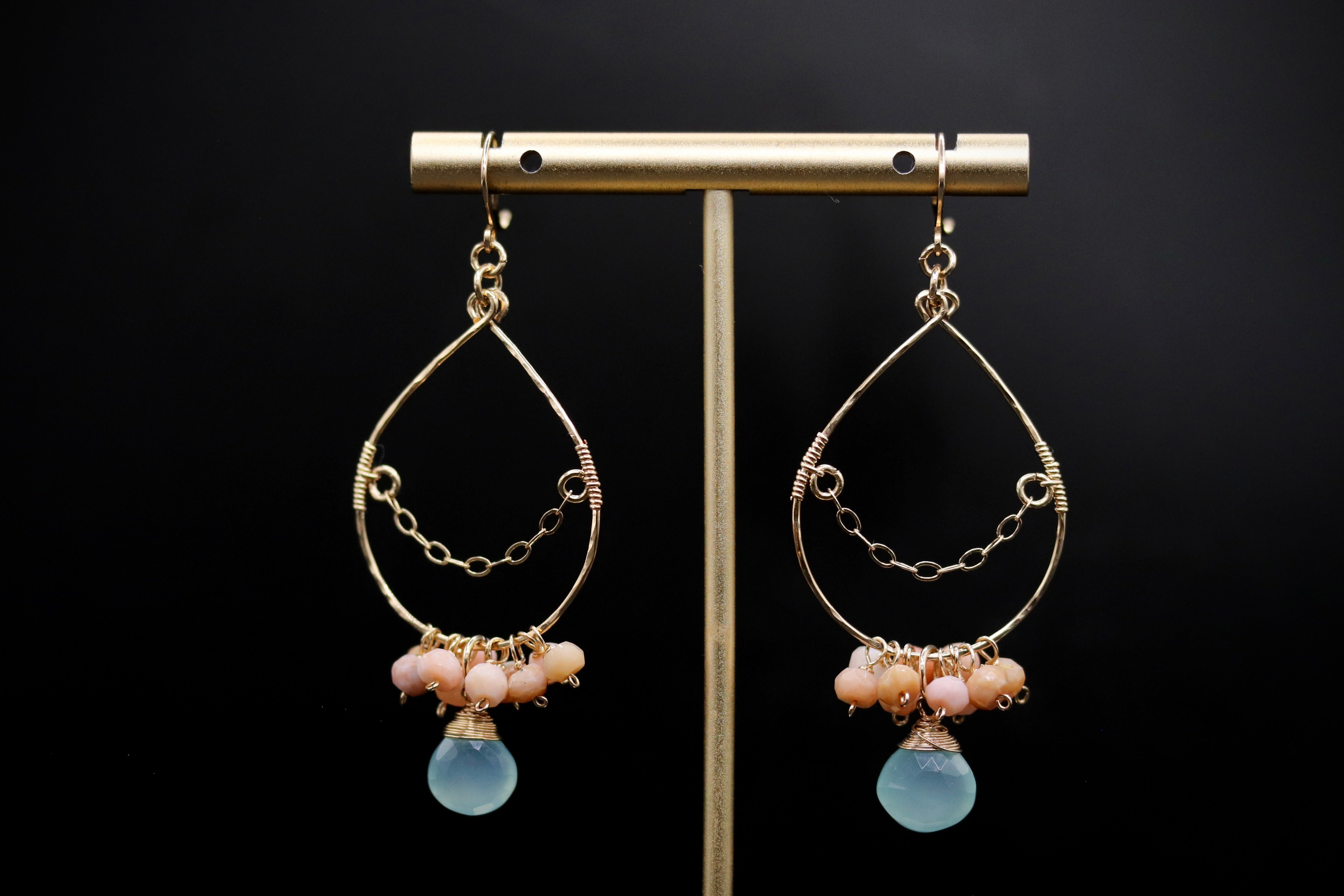 A pair of gold earrings with blue and peach-colored stones hanging from them, displayed on a gold stand against a black background.
