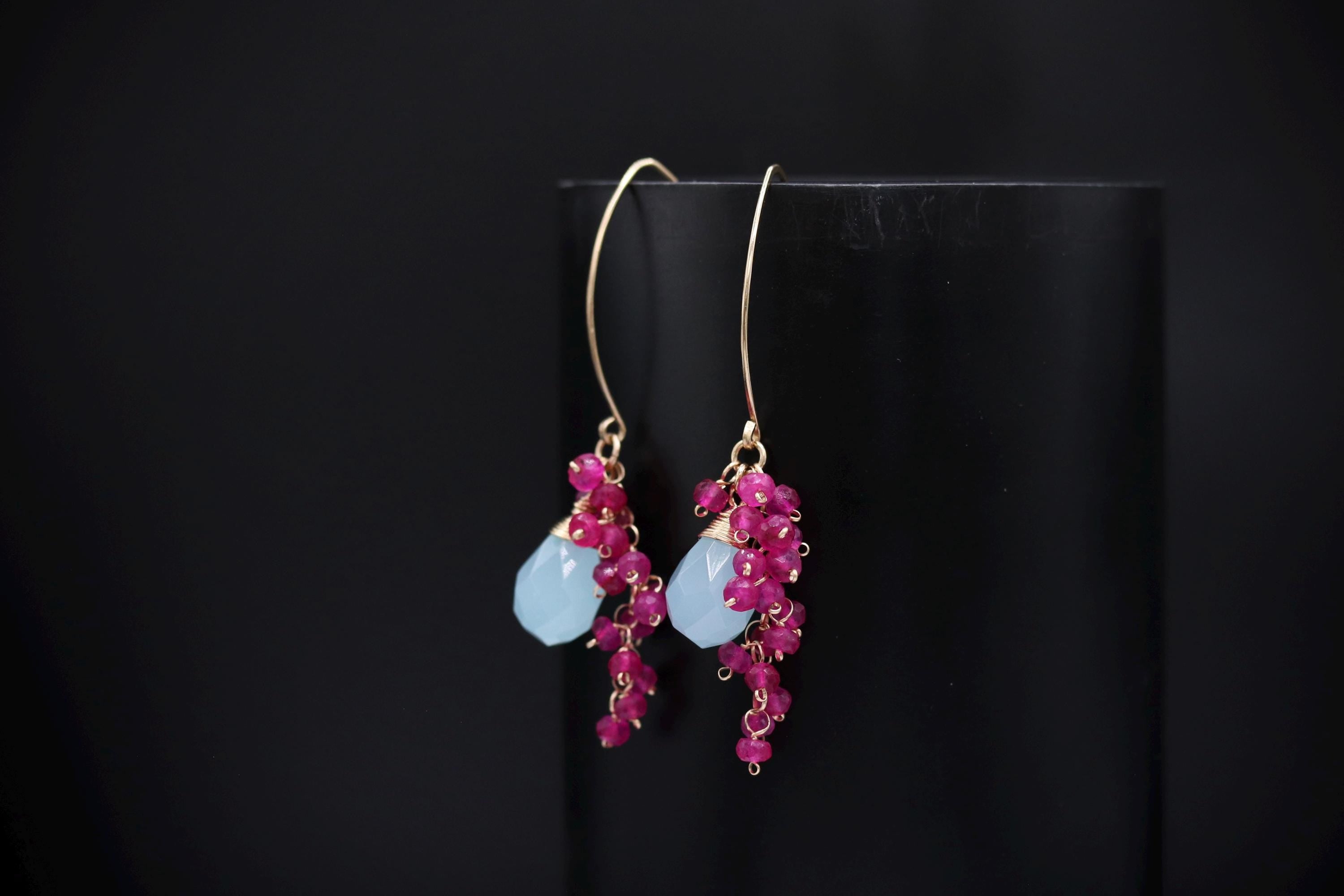 A pair of earrings with blue stones and red gemstones hanging from a black stand against a dark background.