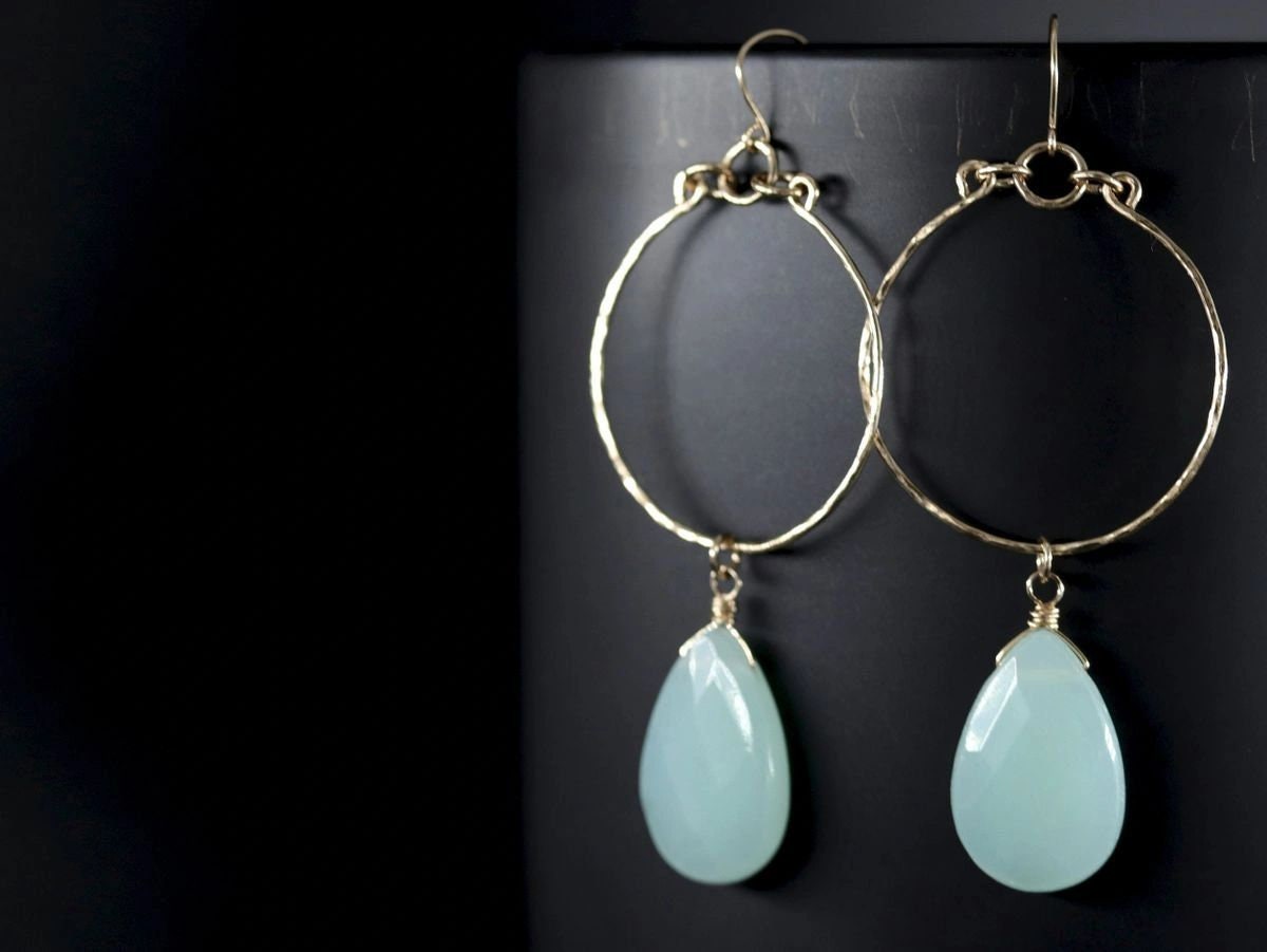 May include: Gold hoop earrings with a teardrop-shaped light blue gemstone dangling from each hoop.