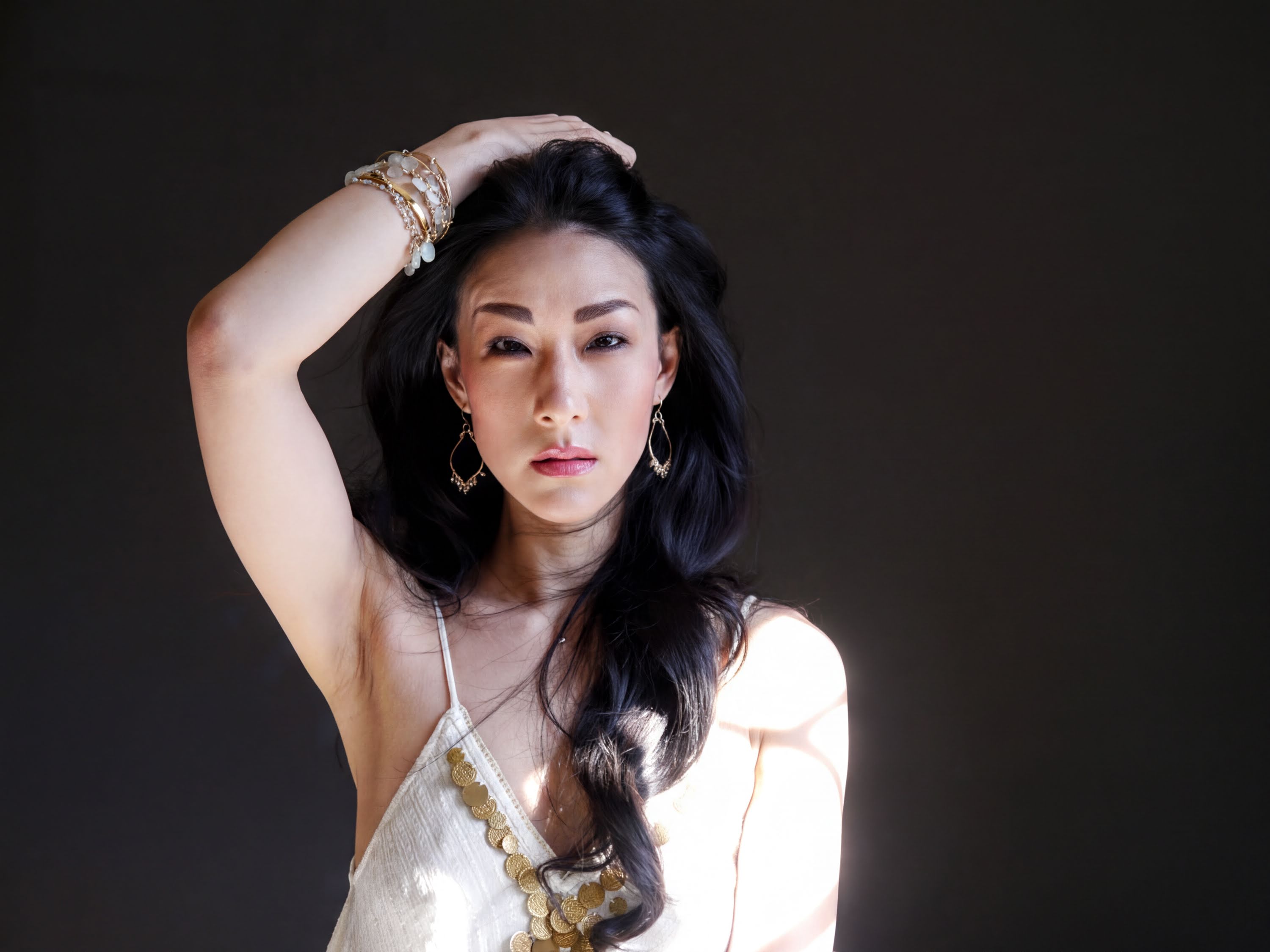 A woman with long dark hair wearing a white dress with gold accents poses with her hand on her head against a dark background.