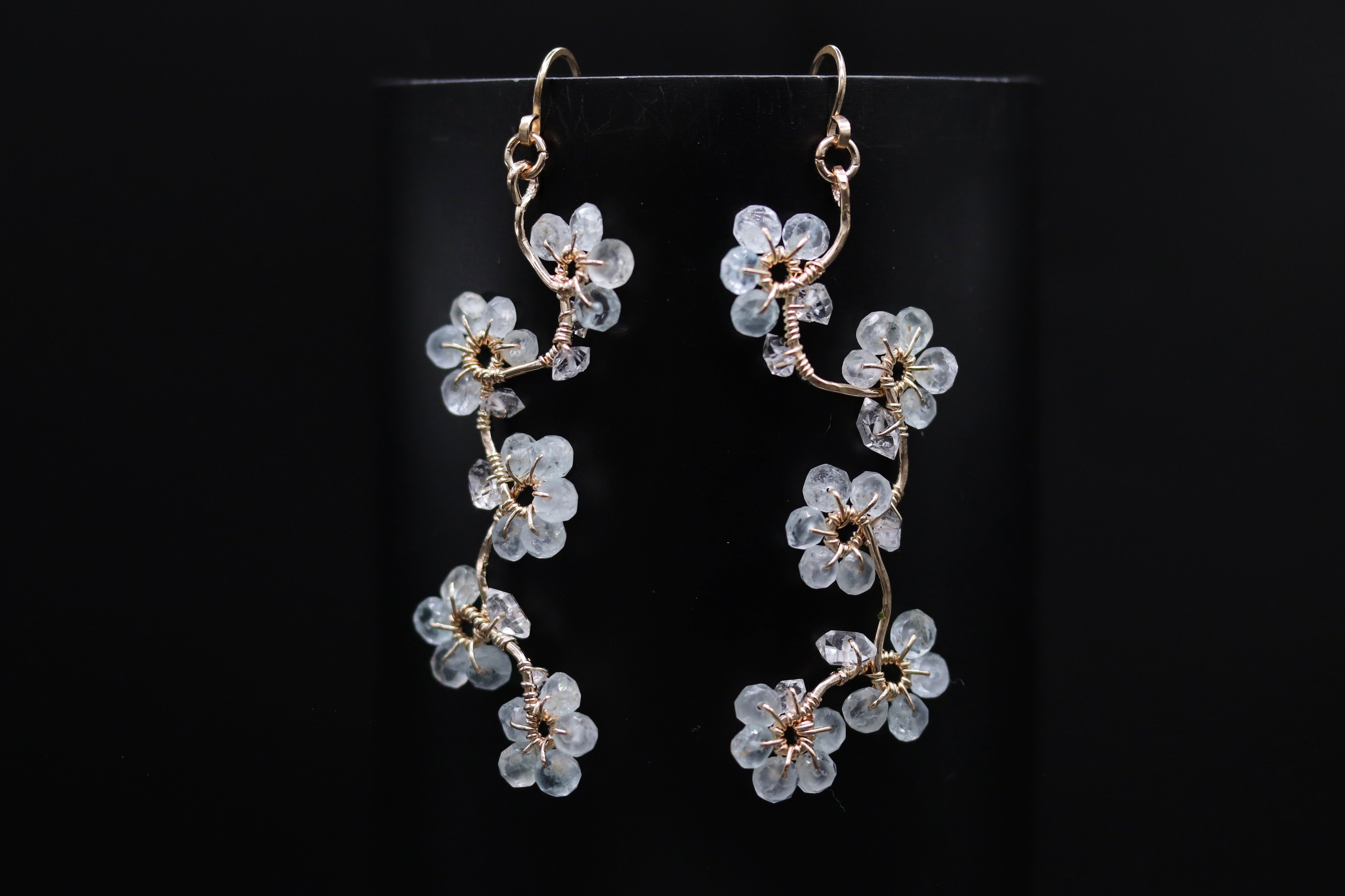 A pair of delicate earrings with white flower-like designs hanging from a thin, curved metal wire against a dark background.