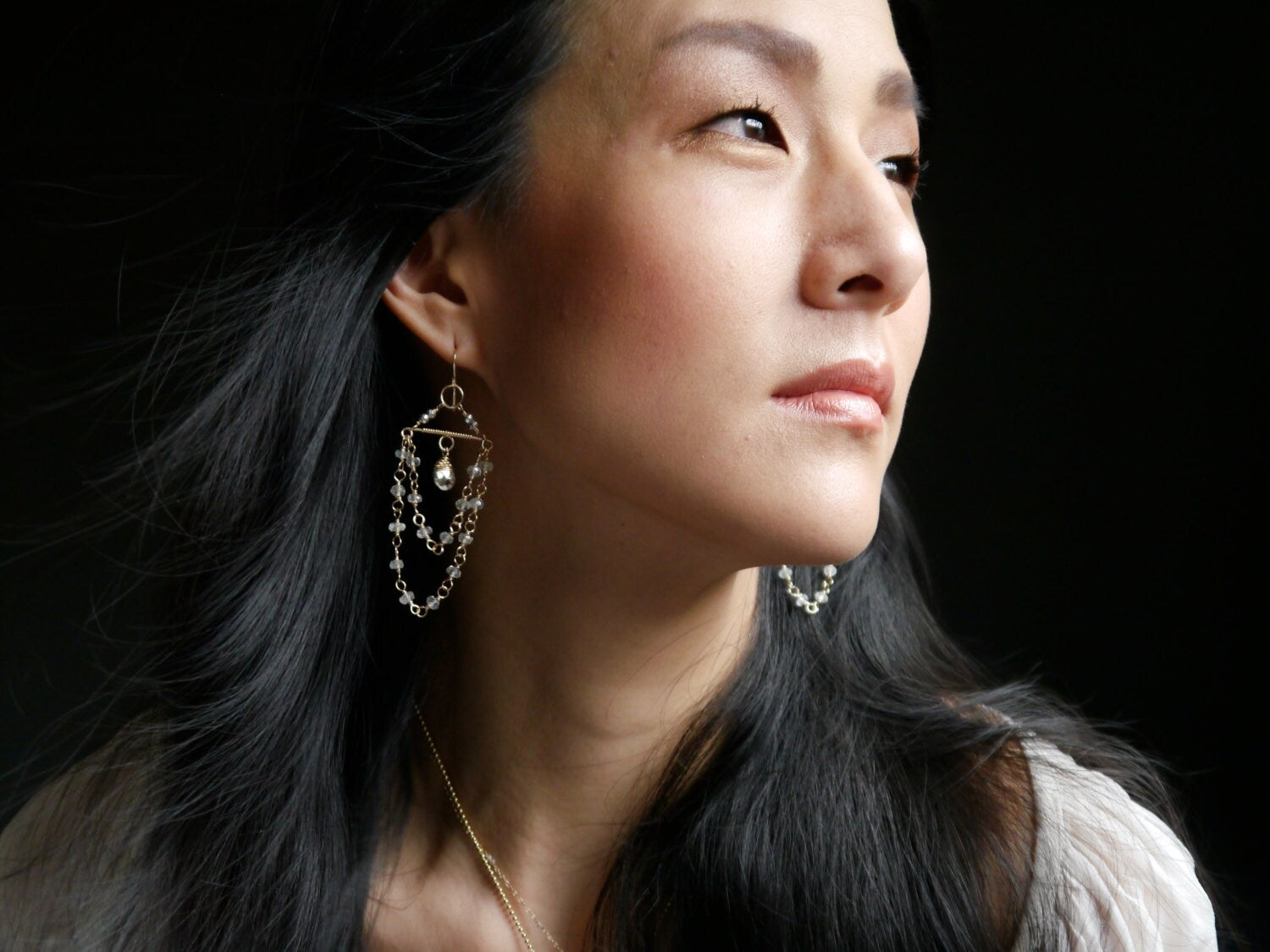 May include: Gold-toned earrings with a layered design. The earrings feature multiple strands of small, clear beads and a central teardrop-shaped charm. The design is geometric, with a horizontal bar at the top and cascading chains.