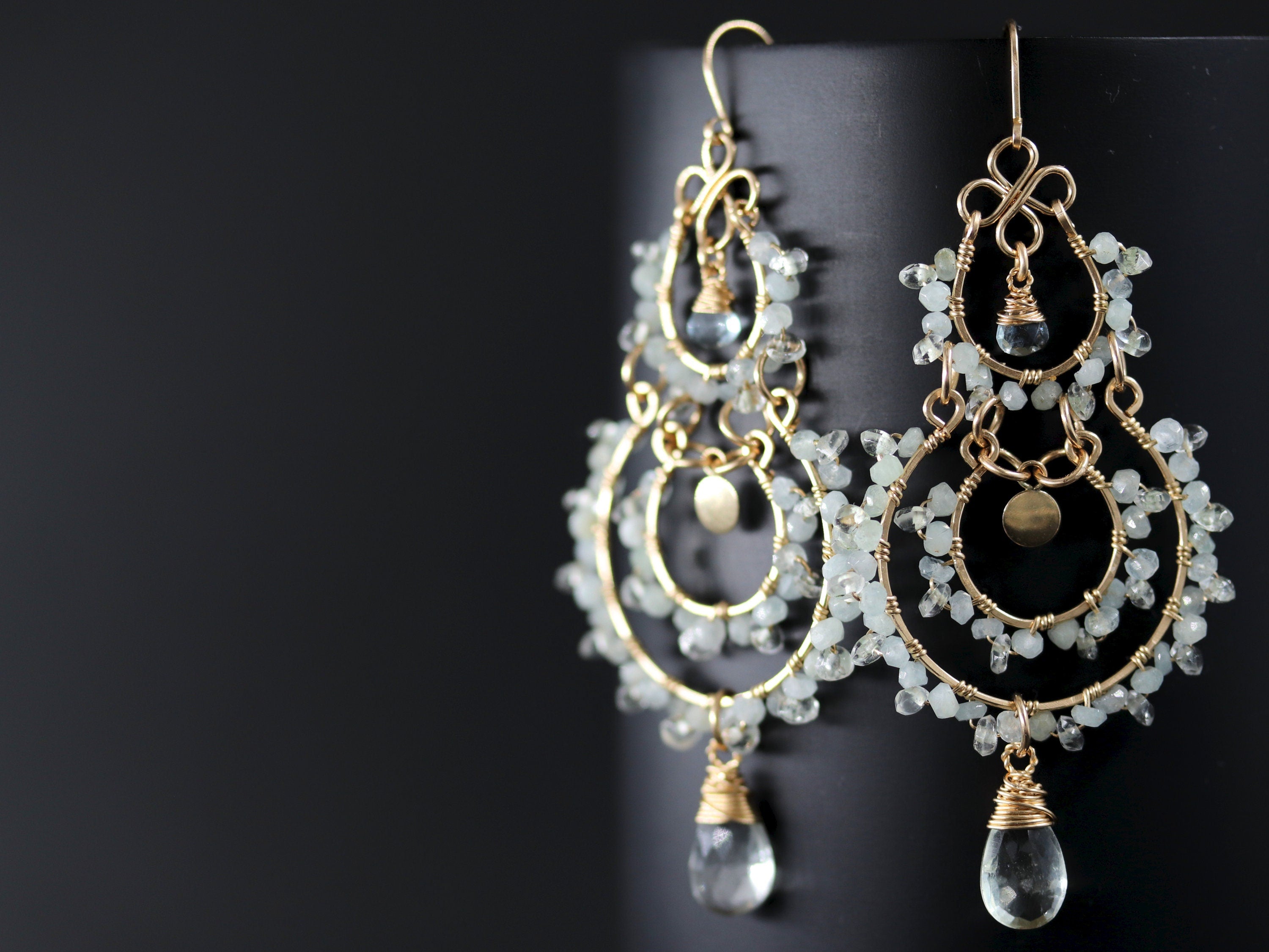 May include: Gold-toned chandelier earrings with multiple tiers of small, pale green beads and teardrop-shaped pendants. The earrings have a delicate, handcrafted appearance, with intricate wirework and small gold accents.