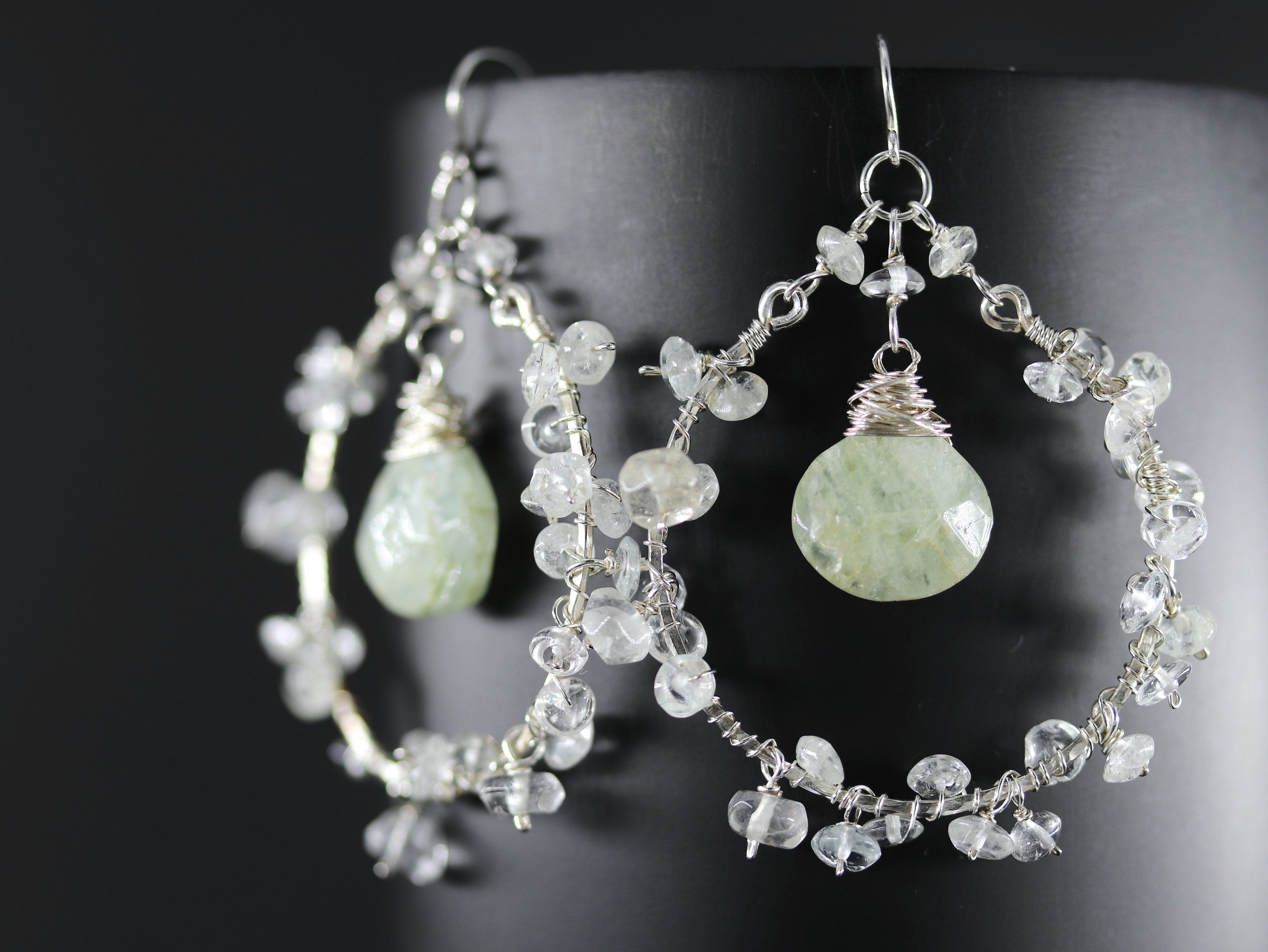 May include: Silver hoop earrings with a light green gemstone dangle and clear crystal accents.