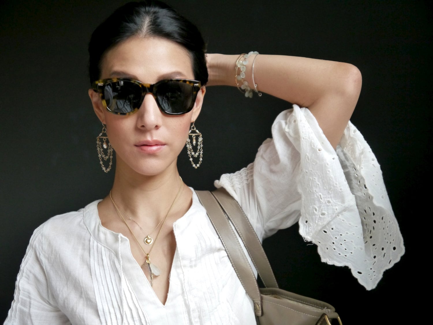 May include: A woman wearing tortoise shell sunglasses, gold earrings, and layered necklaces. She is wearing a white eyelet blouse and has a light brown purse over her shoulder. A bracelet is on her wrist.