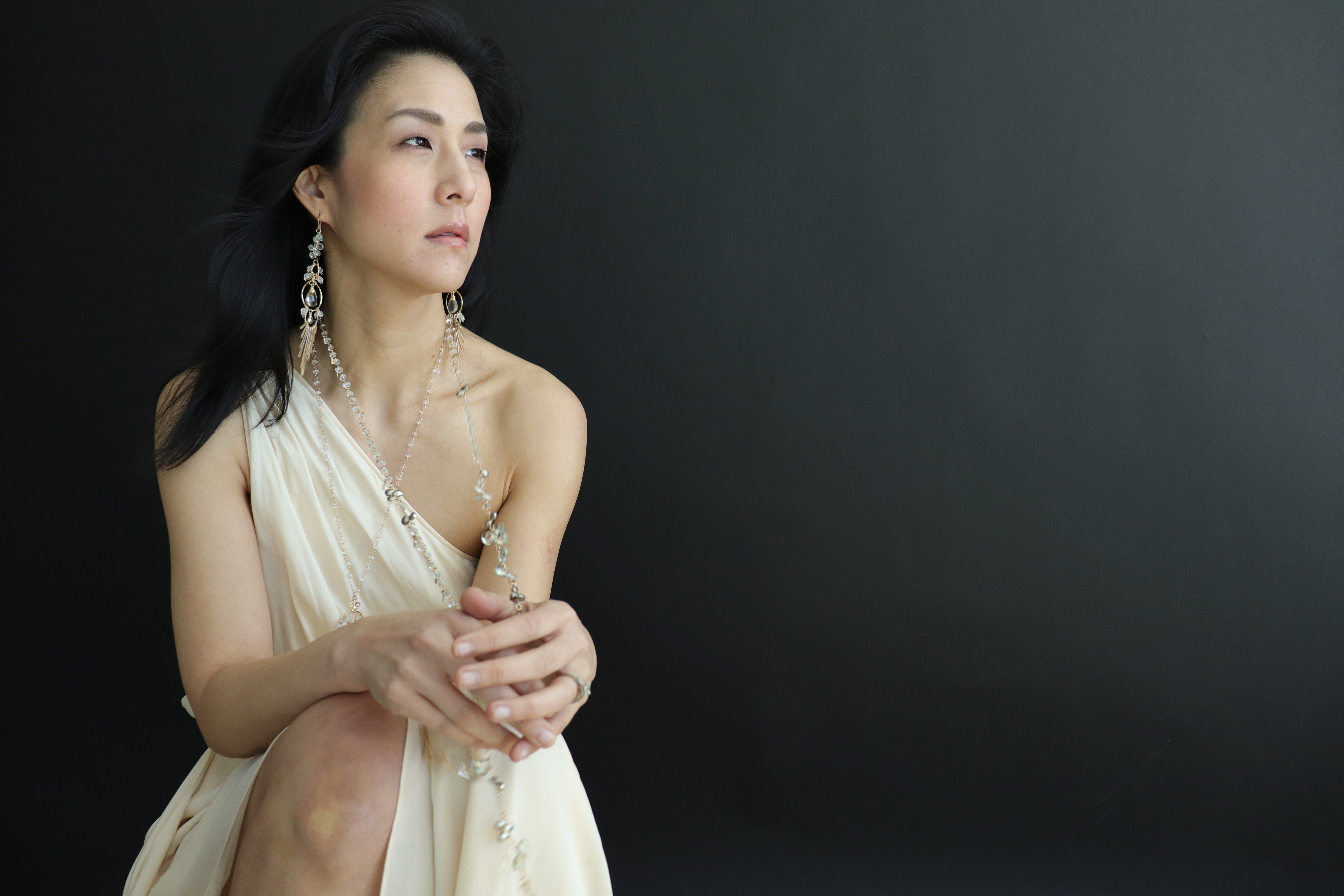A woman in a white dress with long dark hair and large earrings stands against a dark background, gazing off to the side with a thoughtful expression.