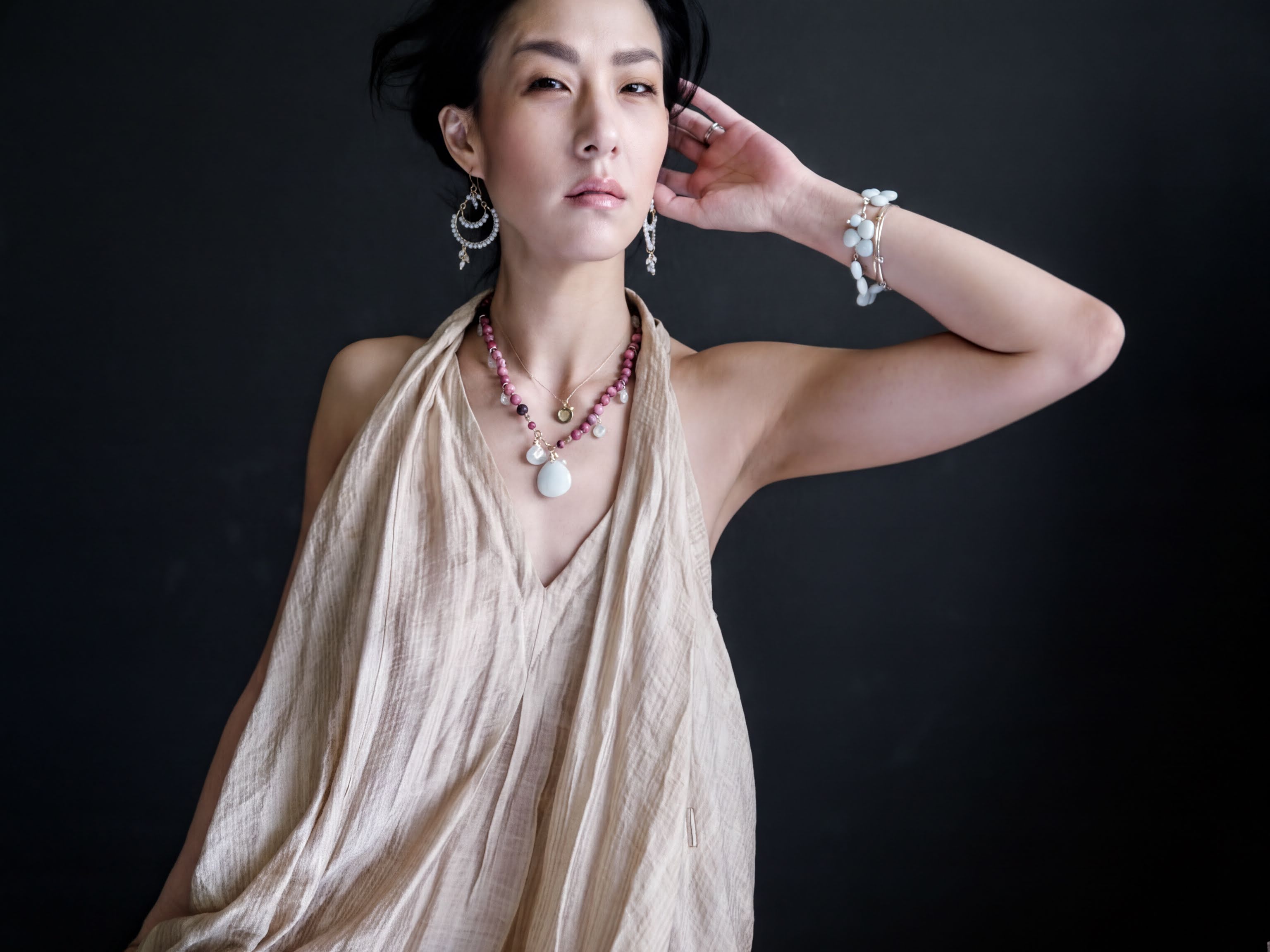 A woman wearing a beige dress and jewelry poses against a dark background.