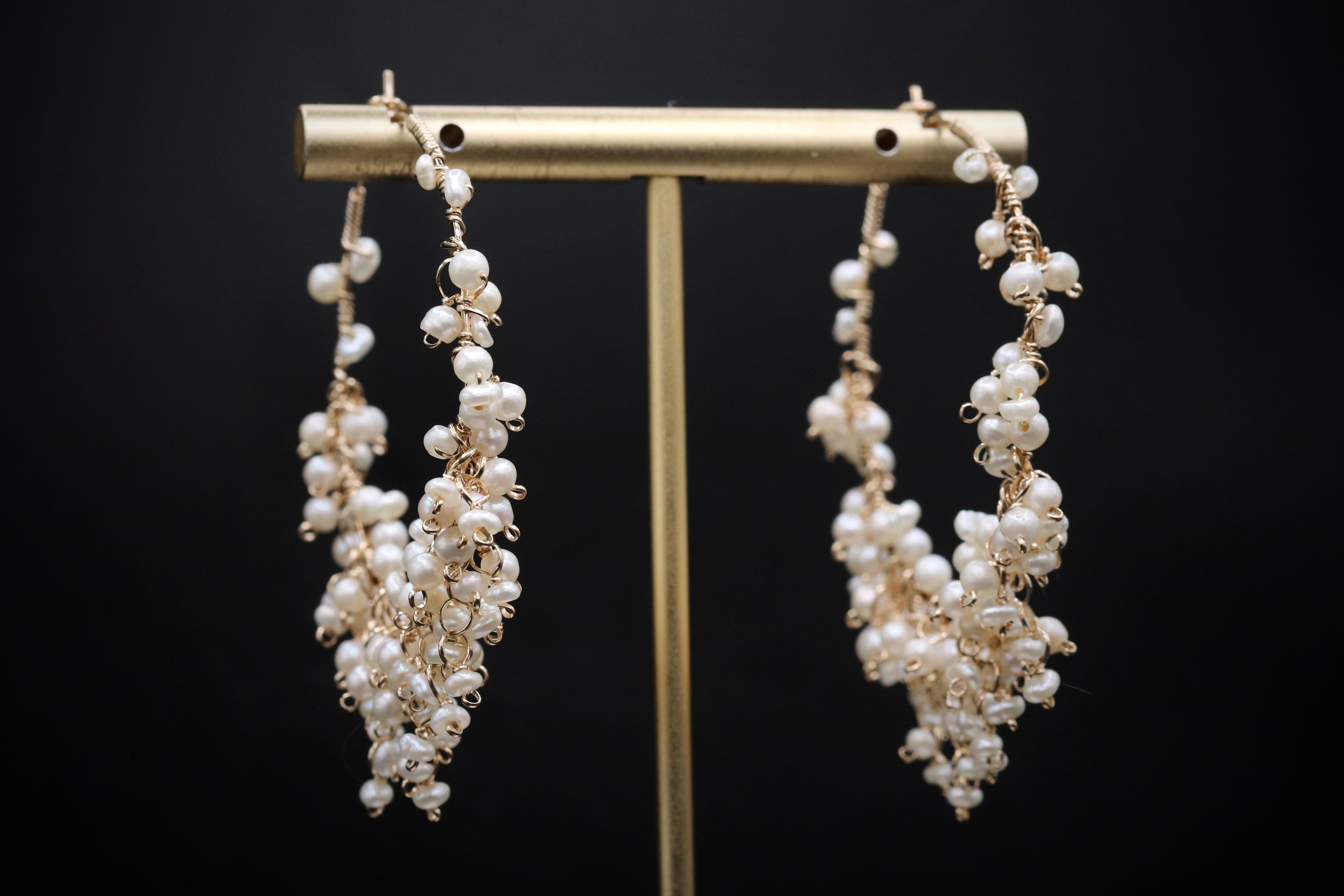 A pair of elegant earrings with numerous small white pearls hanging from a gold-colored metal stand against a black background.