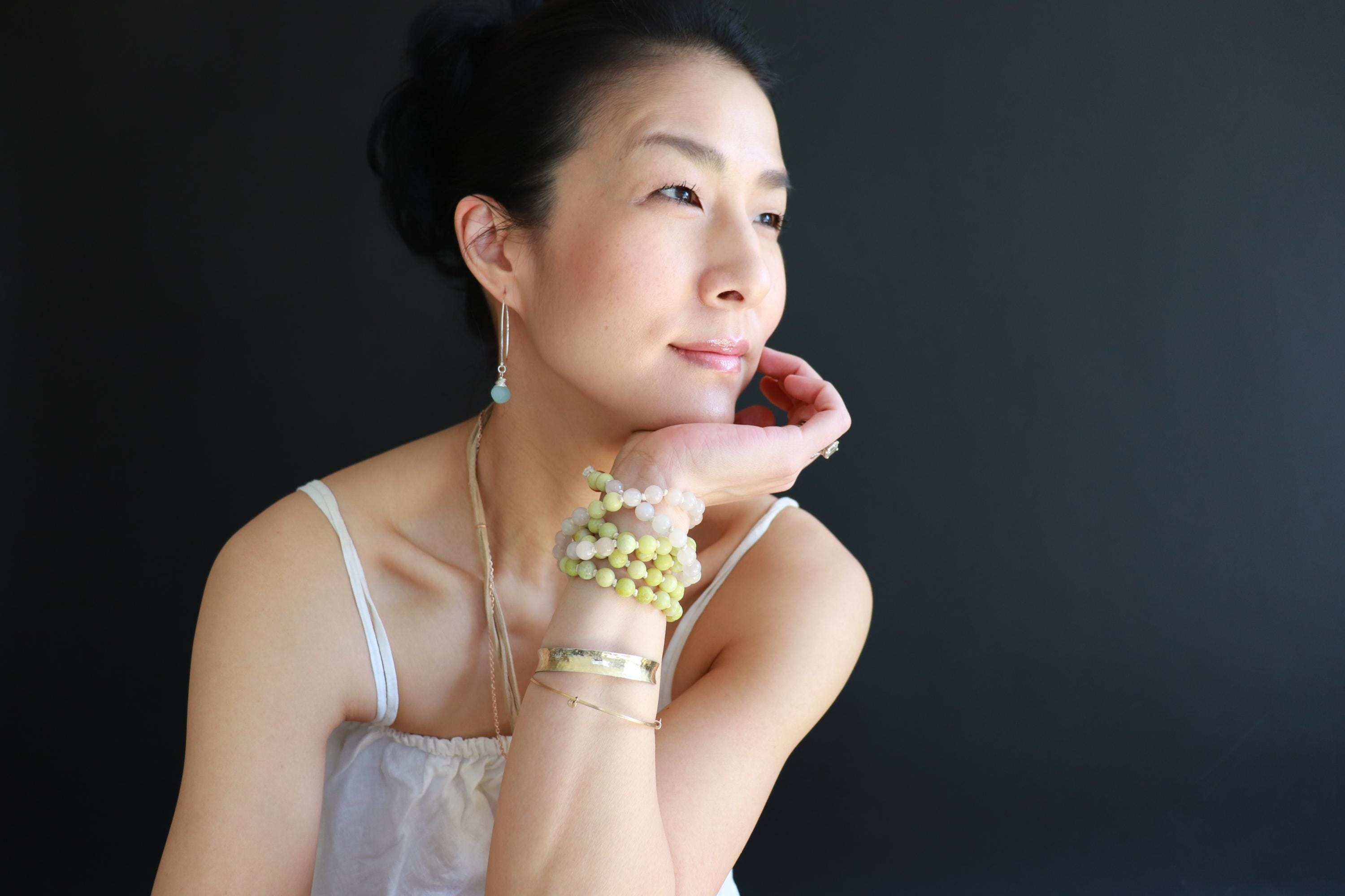 A woman with dark hair and a thoughtful expression, wearing a white tank top and accessorized with bracelets and earrings, against a dark background.